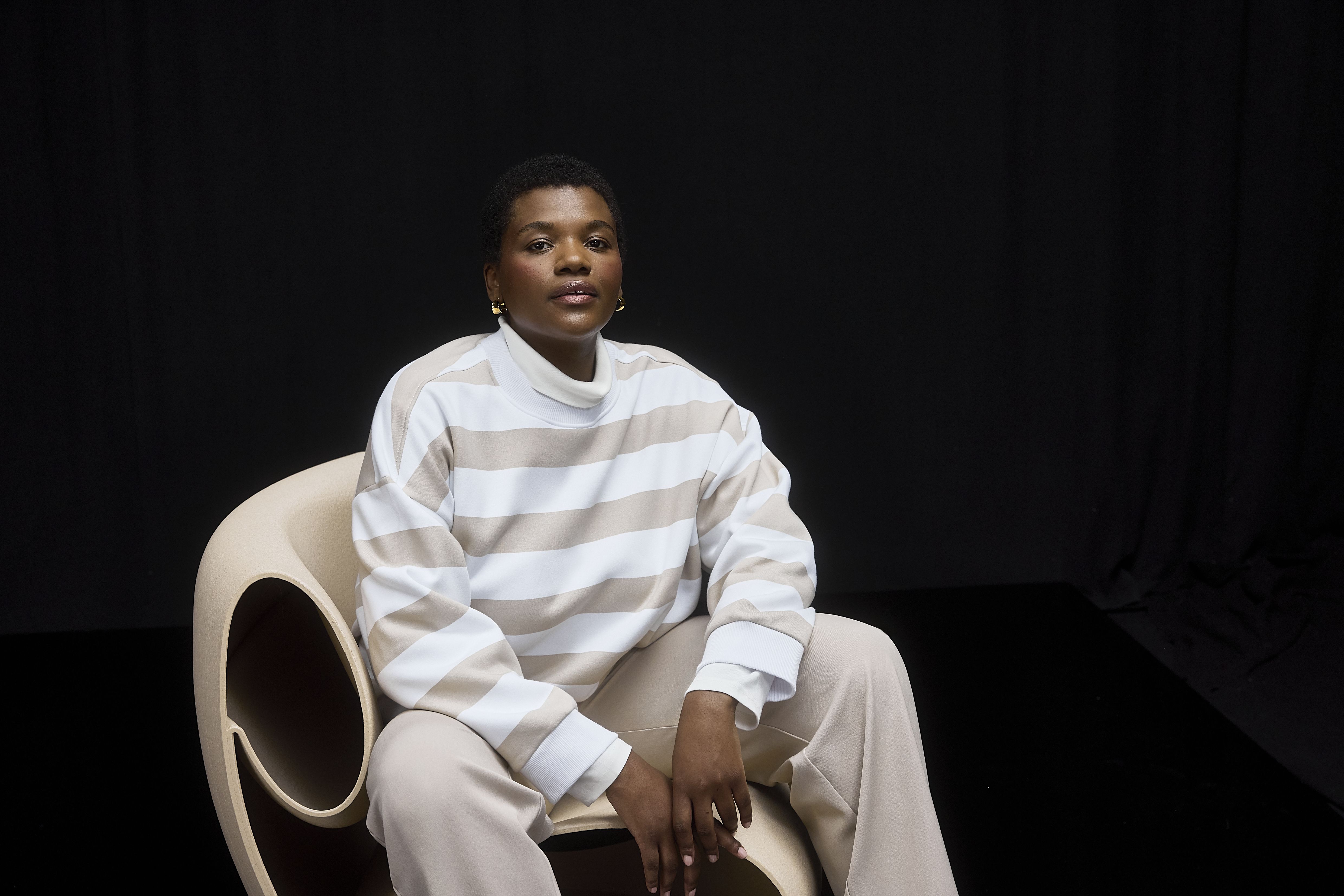 Woman sitting down, wearing beige and white stripped sweatshirt, with beige pants and white sneakers