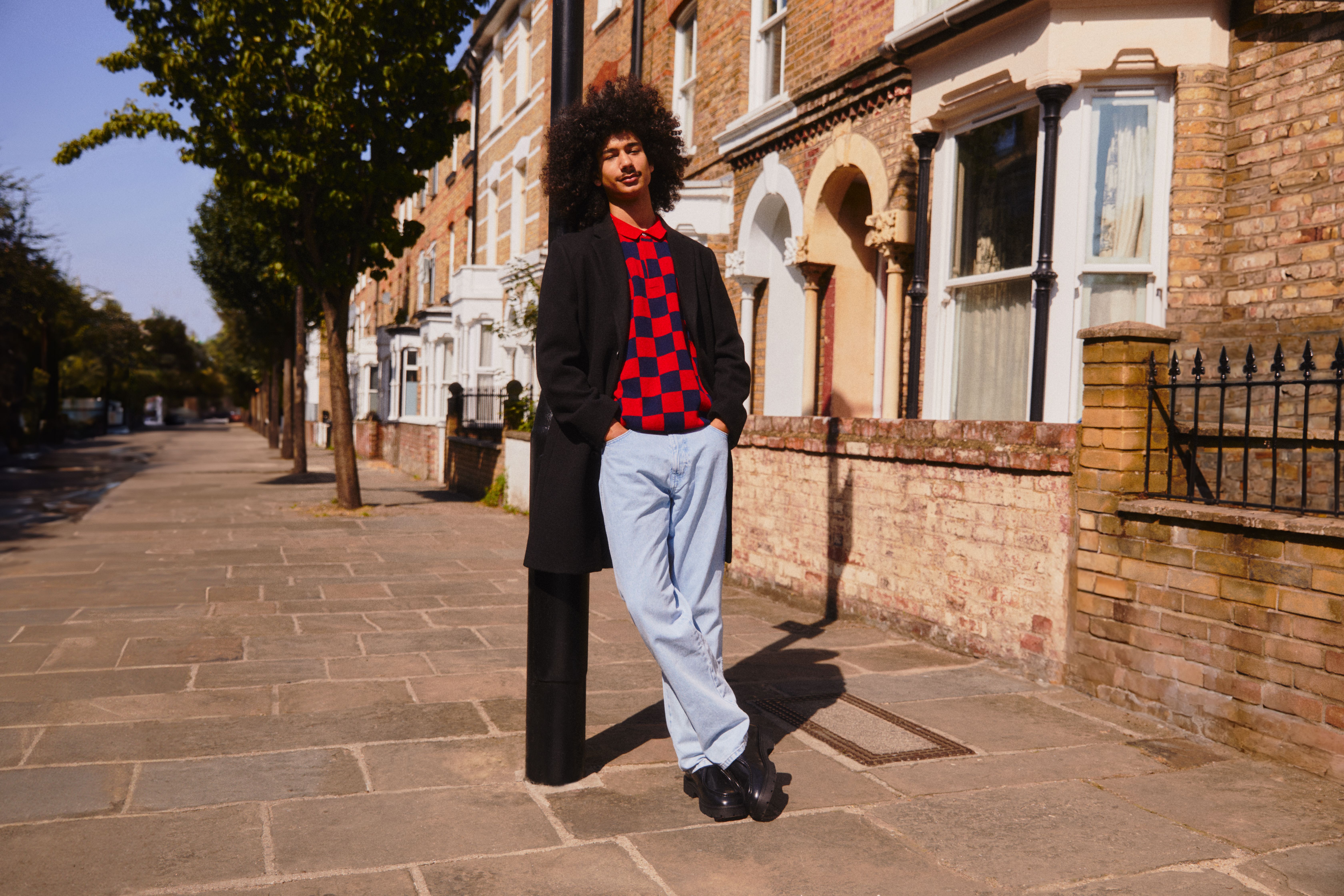 Man leaning on street lamp, wearing vibrant sweater and light jeans that stand out against the background